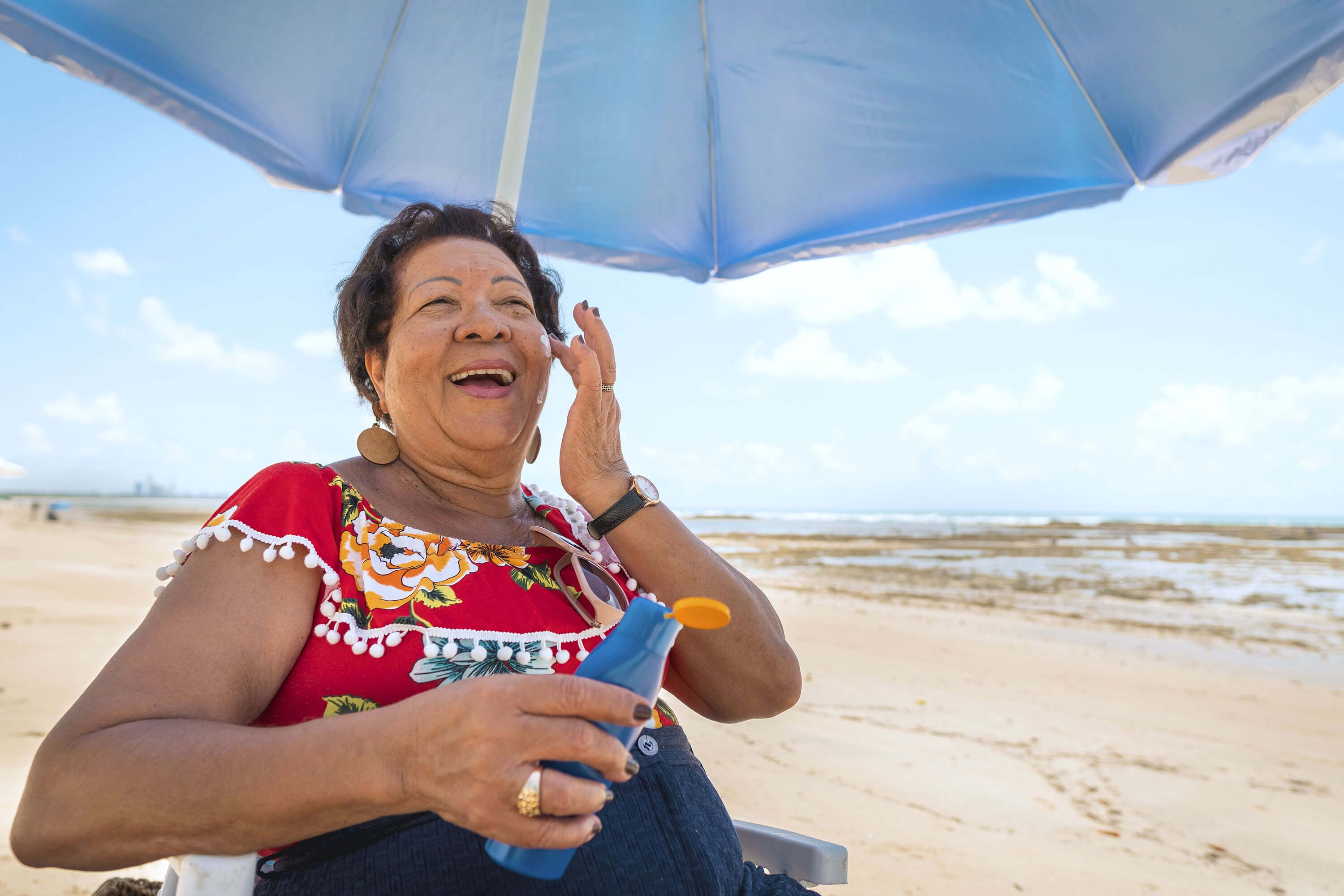 woman smiling on the beach sitting under an umbrella putting sunscreen on her face
