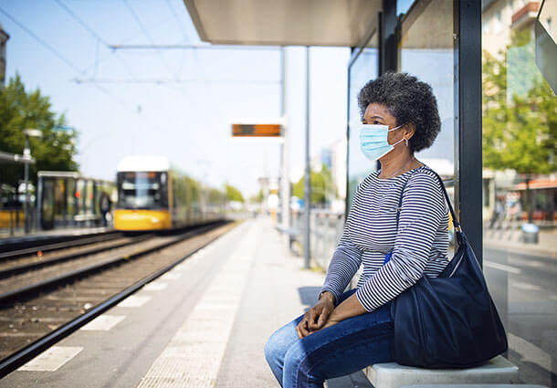 woman wearing a mask sitting on a bench at a train station as a train approaches