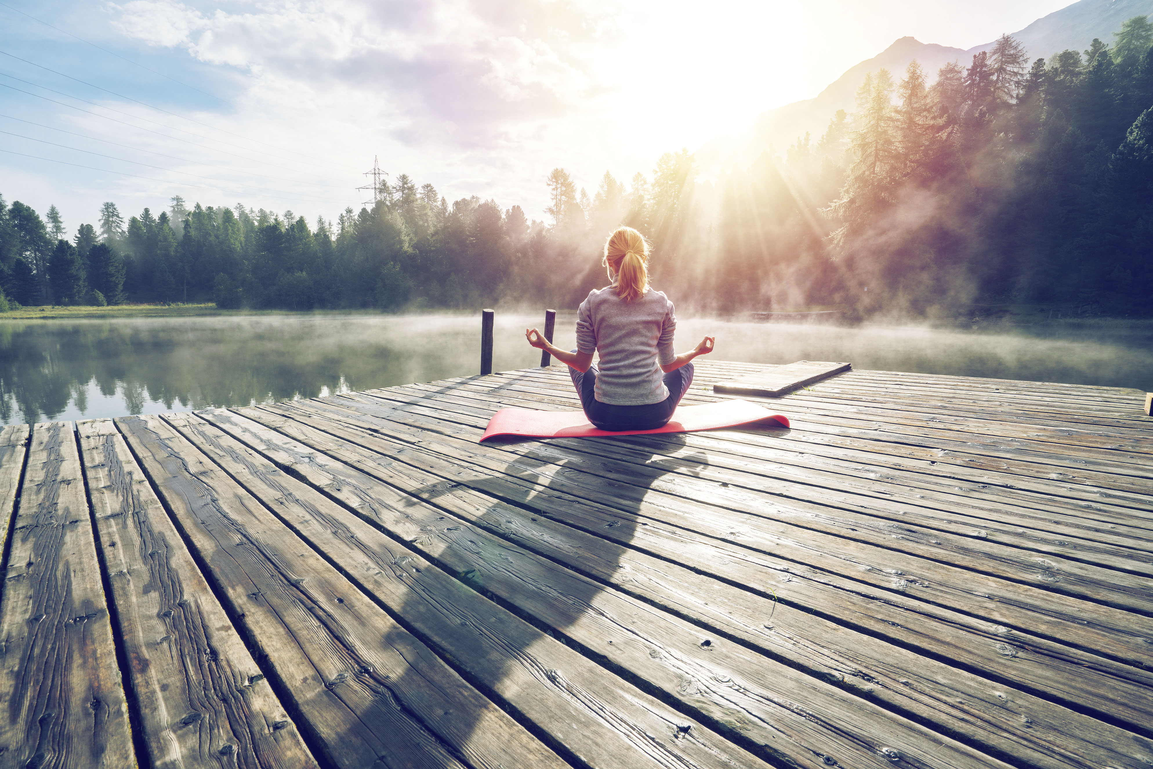 person sitting cross-legged on her yoga mat on a dock overlooking a lake and trees and mountains in the distance