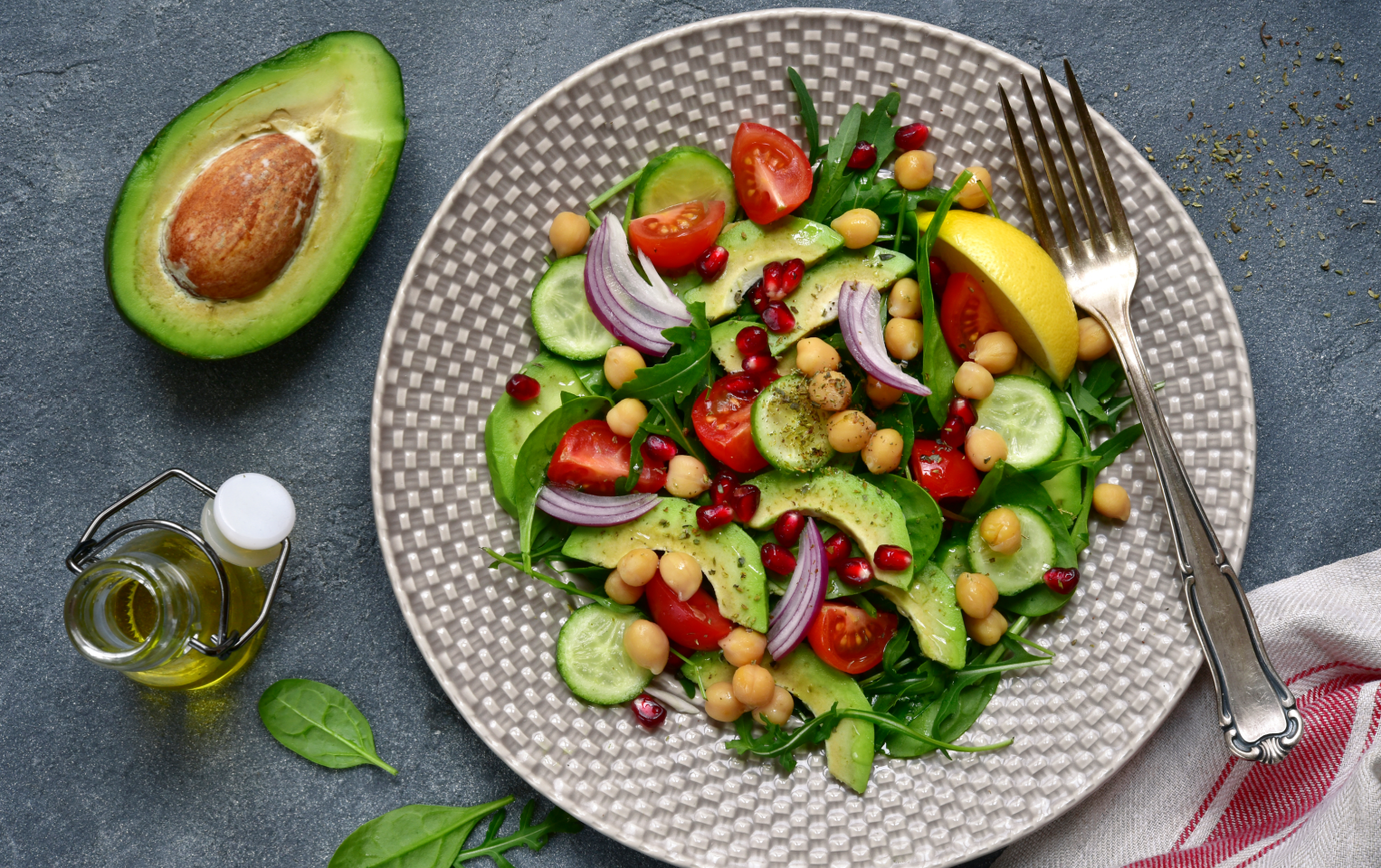 A plate of chickpea, lemon, tomato, and cucumber salad with an avocado and fork on the side placed on a grey table.