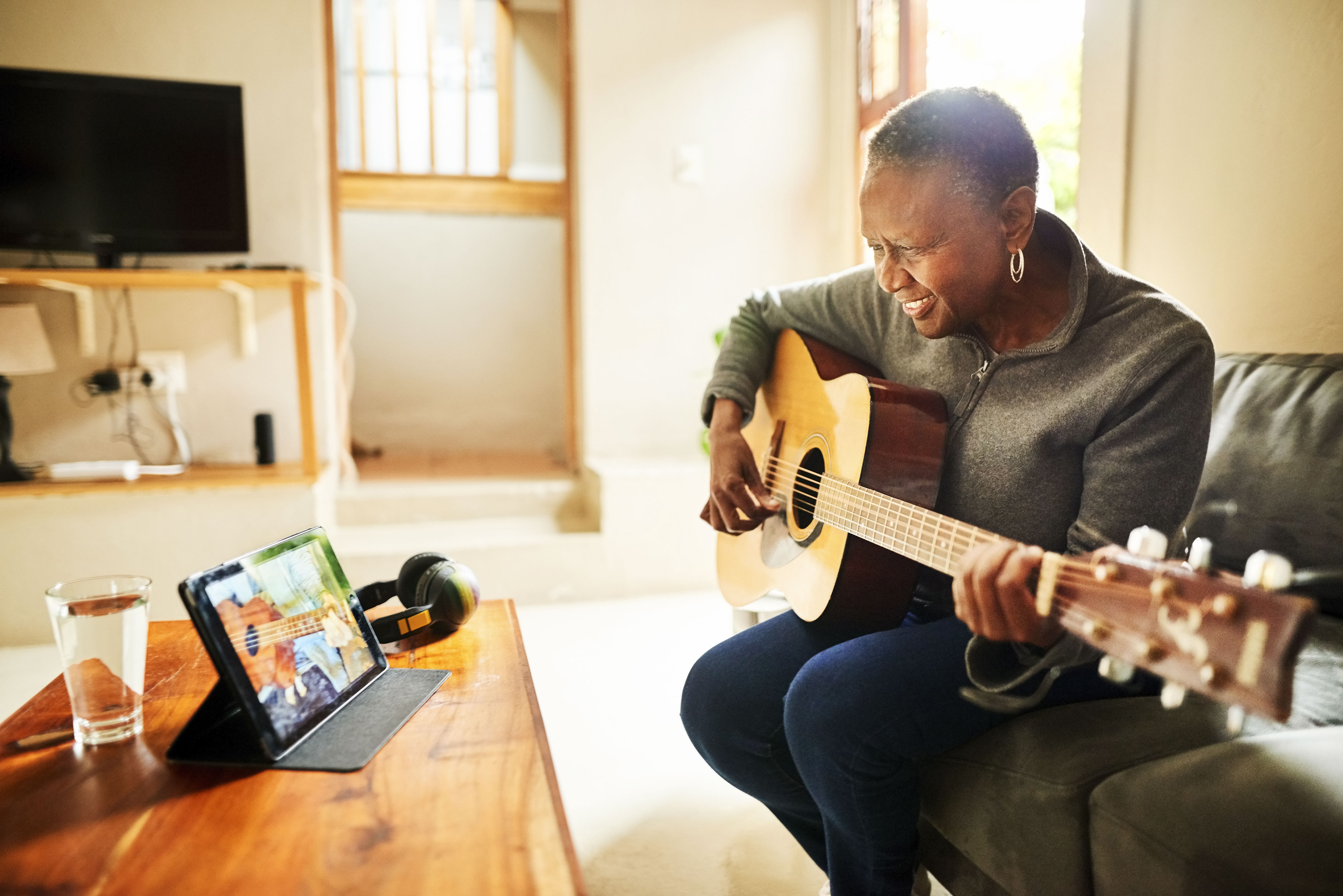 Woman in a green sweater on her couch holding a guitar while watching a guitar tutorial on her iPad