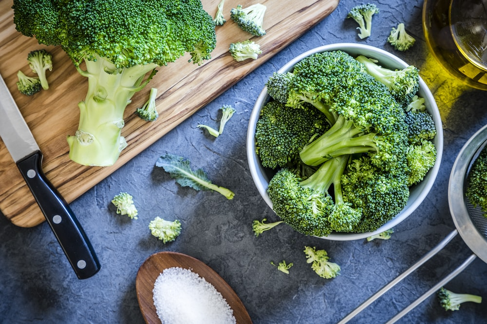 A grey table with a bowl of broccoli, salt, olive oil, and cutting board with a head of broccoli on top paired next to a knife.